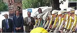 Six cyclists, wearing skin-tight jerseys, sit crouched over their bicycles in preparation to start a race. Men in suits are visible on the far side of the image.