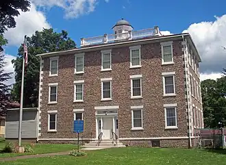 A three-story building faced in small earth-toned rocks with a rounded cupola on top. There is an American flag on a flagpole at left and a blue-and-gold historical marker in front. Above the door are the words "Town Hall".