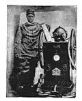 Diver in Standard diving dress posing with diver's pump. His helmet is on top of the pump.