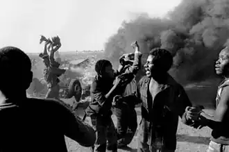 a group of 6 Black South Africans near an overturned car and smoke