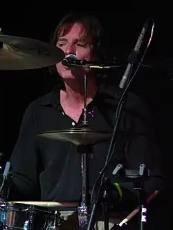 The drummer in a close-up view through the gaps between his drums and cymbals. His face and neck are slightly reddened from his exertion. While drumming, he is singing into the microphone mounted close to his face