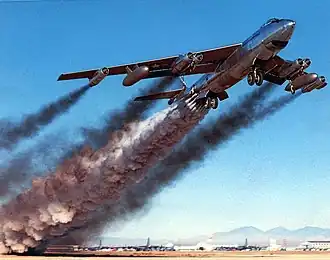 A shiny needle-nosed four-engine jet aircraft takes off. It is trailing an enormous amount of smoke.