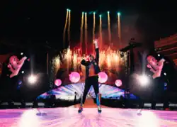 Martin singing with a microphone as the stage behind him release numerous fireworks