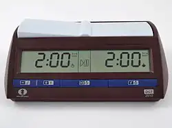 A brown chess clock with blue buttons along the bottom. A digital display shows the time remaining for each side