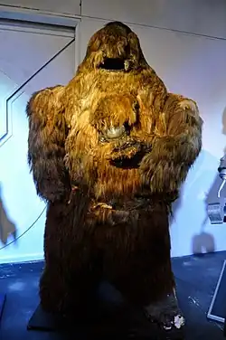 An image depicting a tall, furry, humanoid costume. Its arms are crossed over its stomach.