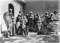 Woman in 19th century British dress stood amongst people in Afghanistan dress. She is positioned beside a doorway, and is examining the eye of a child.