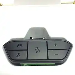 Xbox One Stereo Headset Adapter, a black plastic attachment that plugs into the bottom of the Xbox One Wireless Controller and provides a standard 3.5&nbsp;mm stereo headset jack with microphone contact. The adapter itself is roughly trapezoidal and has five buttons on its face; two buttons on the left to balance audio output between in-game chat (bottom) and game (top); a large center button to mute the chat microphone; and two buttons on the right to increase (top) or decrease (bottom) overall volume.
