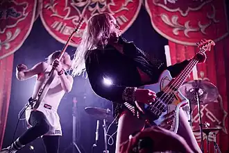 Angel (foreground) and Lulu (background) of Frost Children, playing the electric guitar during a climactic moment of a song. Illuminated by stage lights and backed by theatre curtains.