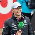 George Russell wearing a silver baseball cap on his head, a black coat with sponsors logos and he is holding a microphone in his right hand