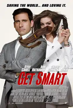 Maxwell Smart wearing suit, his tie blown to the side, covering the face of Agent 99 who is standing behind him wearing a white jacket and holding a gun.