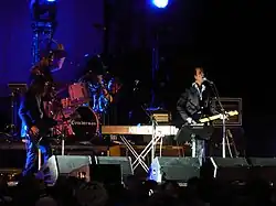 A rock band performing live onstage. On the left, a man is playing a bass guitar and a man is playing the drums. A man is singing and playing guitar in the center-right.