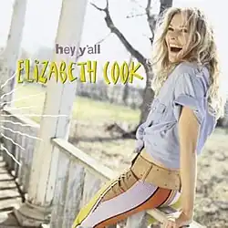 An image of a blonde woman laughing while leaning against the railing of a porch. She is wearing a blue shirt and white-orange striped pants. The words "hey y'all" appear in a purple font while "ELIZABETH COOK" is included below it in a yellow font.