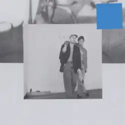 A black-and-white photograph of two men with one's arm around the other set against a larger indistinct photograph, with a blue square in the top-right of the image.