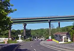 A pair of bridges pass from left to right on tall supports that tower over the other structures in the area, such as telephone poles and single-story buildings. The two-lane NY&nbsp;22 passes underneath the bridge and proceeds into the background.