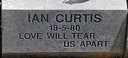A grey stone with "Ian Curtis, 18-5-80, Love Will Tear Us Apart" carved into it in block letters