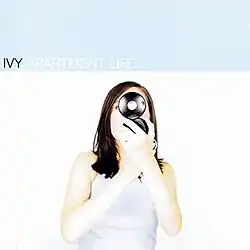 Dominique Durand appears facing the camera but holding a makeup compact up to her head, covering the majority of her face.