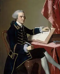 Full-length portrait of a young man seated at a table. He wears a finely tailored dark suit, knee breeches with white stockings, and a wig in the style of an English gentleman. He holds a quill pen in his right hand, and is turning the pages of a large book with the other hand.