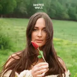 A woman with very long brunet hair holds up a flower against a bucolic green backdrop.