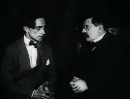Thin, intense young man and mustachioed older one, looking at each other
