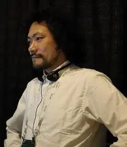 Portrait photo of a Japanese man with a black beard and long frazzled hair facing left.