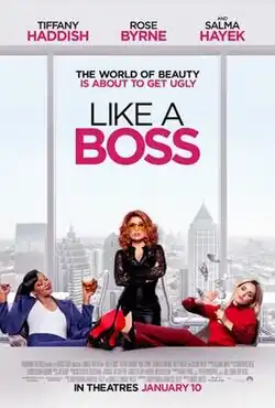 A red-haired woman wearing a black outfit stands with her arms crossed at a boardroom table, behind her is view of the city. In front sit two women with their feet up on the table, one dressed in blue, the other in red.