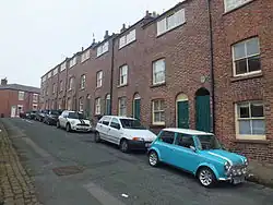 A row of nine identical brick cottages, with a green door and rising sash window on the ground floor, and a single rising sash window on the first floor. Above, built into the roof is a wide window, twelve panes long and four panes high. The cottages are built directly on the street, in front of one is a turquoise and white T registered Mini.