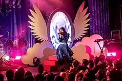 Mica Tenenbaum singing with a microphone in a blue outfit in front of a mirror-shaped screen.