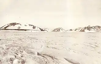 A mountain with a rounded summit and snowy slopes, with smaller hills to its right, rising from flat snow-covered ground