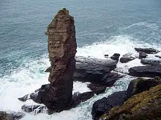 A tall sea stack consisting of a layered sedimentary rock is situated off a rocky coast and sits amid breaking waves and foamy waters.