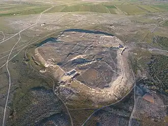 Aerial view of a massive hill, on which the ruins of walls and buildings can be seen, surrounded by wilderness