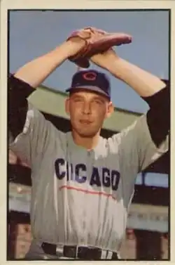 A man in a white baseball uniform with dark sleeves and a dark cap holds his hands together over his head in his glove