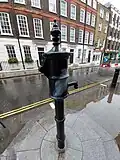 An old-fashioned water pump, painted black, with its handle removed, standing on a stone pedestal two steps above the sidewalk
