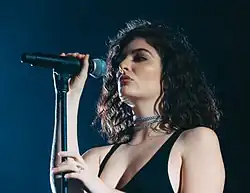A young Caucasian woman with shoulder-length, curly, dark hair wearing two choker chains, a necklace, a revealing black dress and a ring holds a stand-mounted microphone.