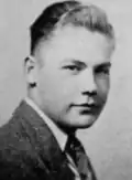 A black and white photo of a man oriented towards the right of the image, turned towards the camera, smiling slightly