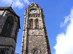 The view up a huge clock tower from the ground upwards. The ornate, angular tower is made of stone and appears a light-taupe colour. It fills the middle part of the image. The top of the tower has a large beige clockface, and above that, a stone spire. In the background is the sky which appears mostly as vivid azure but partly with white clouds.