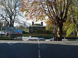 A photograph of the T junction of a road taken from the stalk of the T. A river runs across the top of the T and a small brick cottage can be seen on the far bank.