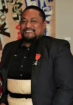a man with short black hair, a tidy beard and moustache smiles directly at the camera. he has a black jacket with a medal and a black shirt. He is also wearing a fine mat woven from natural fibres