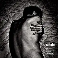 A stark black and white image of a man lying on a mattress. His back is facing the camera and his face is obscured. His hand is resting on his back as if his arms are wrapped around him.