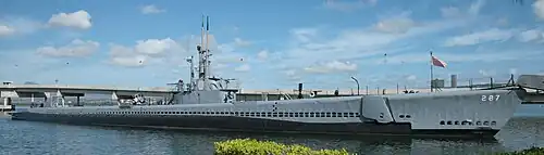 A submarine, long, sleek and painted grey at anchor. It flies a red and white striped ensign.