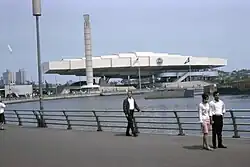 A flat, white, rectangular, elongated building and a tall, cylindrical column on the far shore of a small lake.