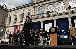 A woman in an American Navy uniform stands before a microphone with several people behind her on stage. The stage is in front of a building with a sign that reads "#27!".